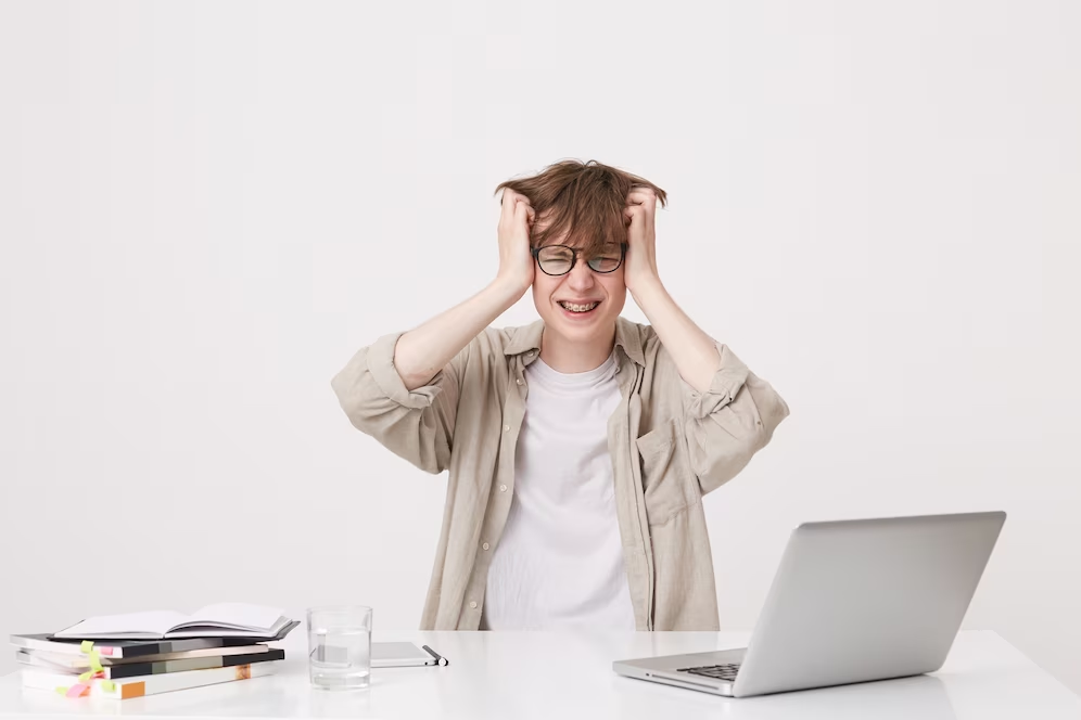 Frustrated man with hands on head at a table with books, laptop, and water.