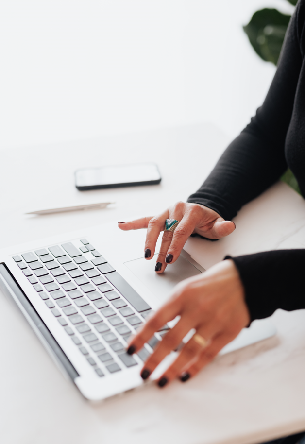 the girl is sitting at the computer, there is a phone and a pen on the table