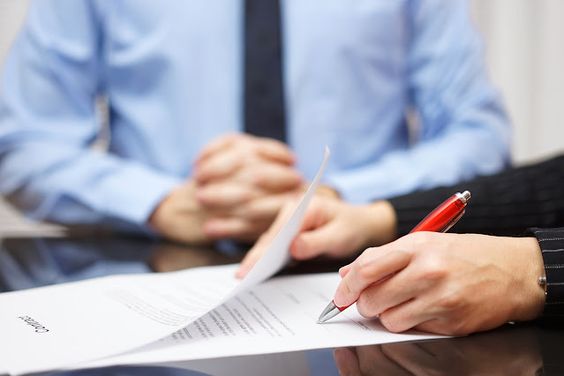 a person concludes an agreement by signing documents, and another person watches with folded hands on the table