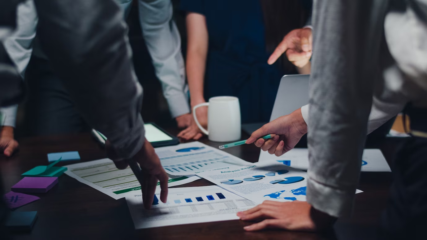 Photo of a group of people gathered around a table, discussing documents containing graphs and pie charts