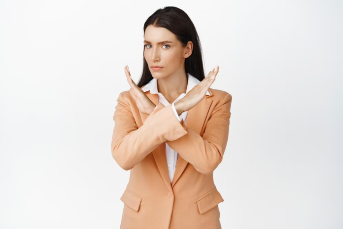 A woman wearing a brown coat, crossing her arms in a 'no' gesture.