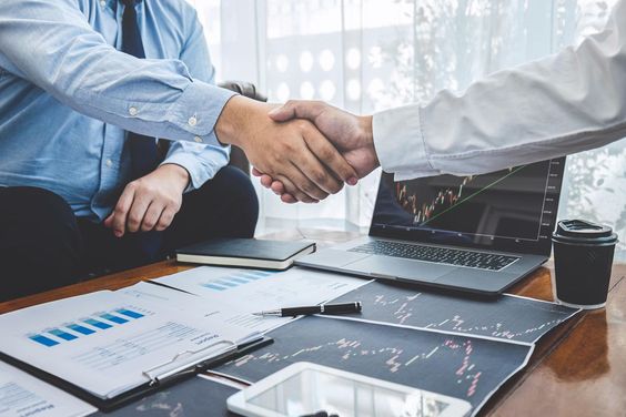 two men make a deal, shaking hands, a laptop, papers, a pen, and coffee are on the table